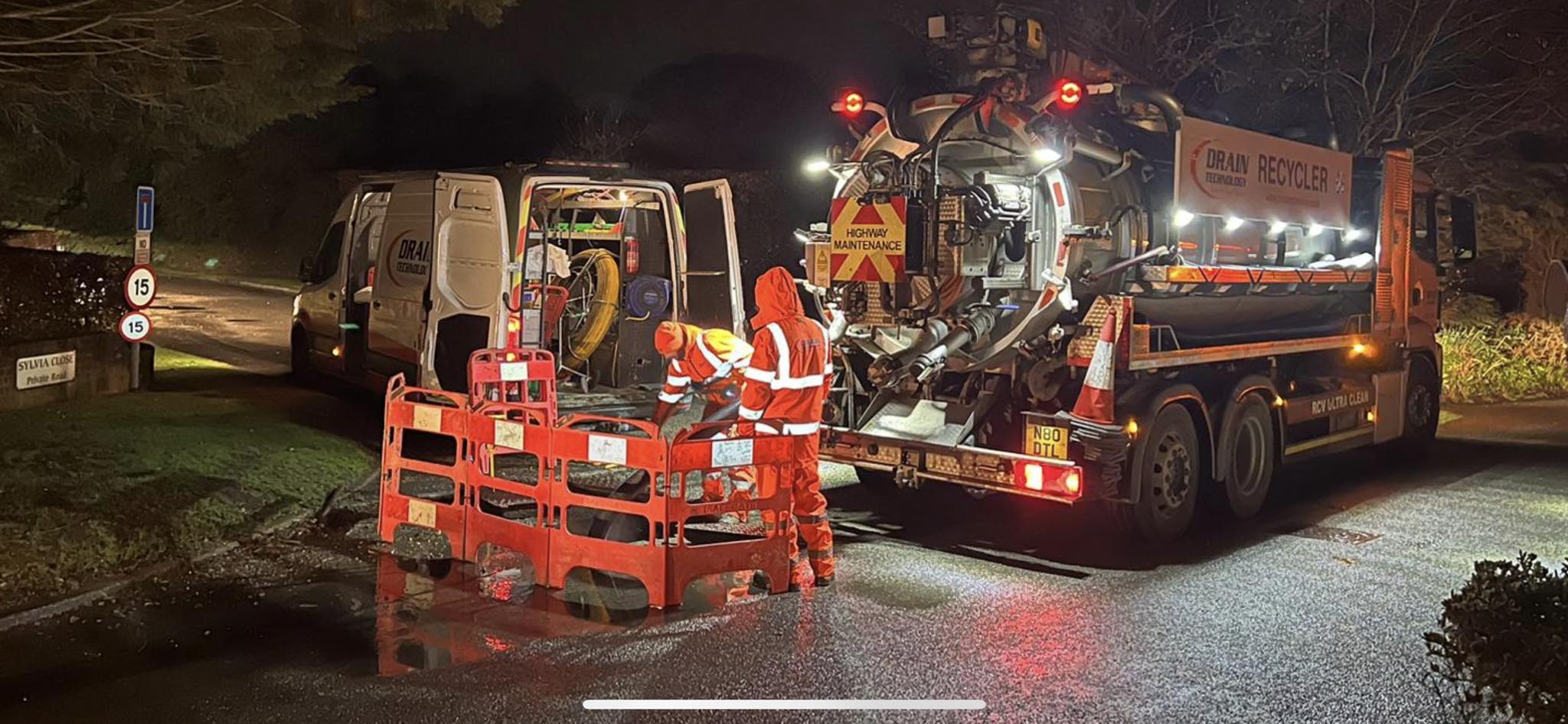 On a crossroads in the middle of the night, a large drain technology recycler vehicle and a large drain technology van are being used. Two drain technology employees wearing hi-vis are standing surrounding the main point of the site which is corned off with construction fencing.