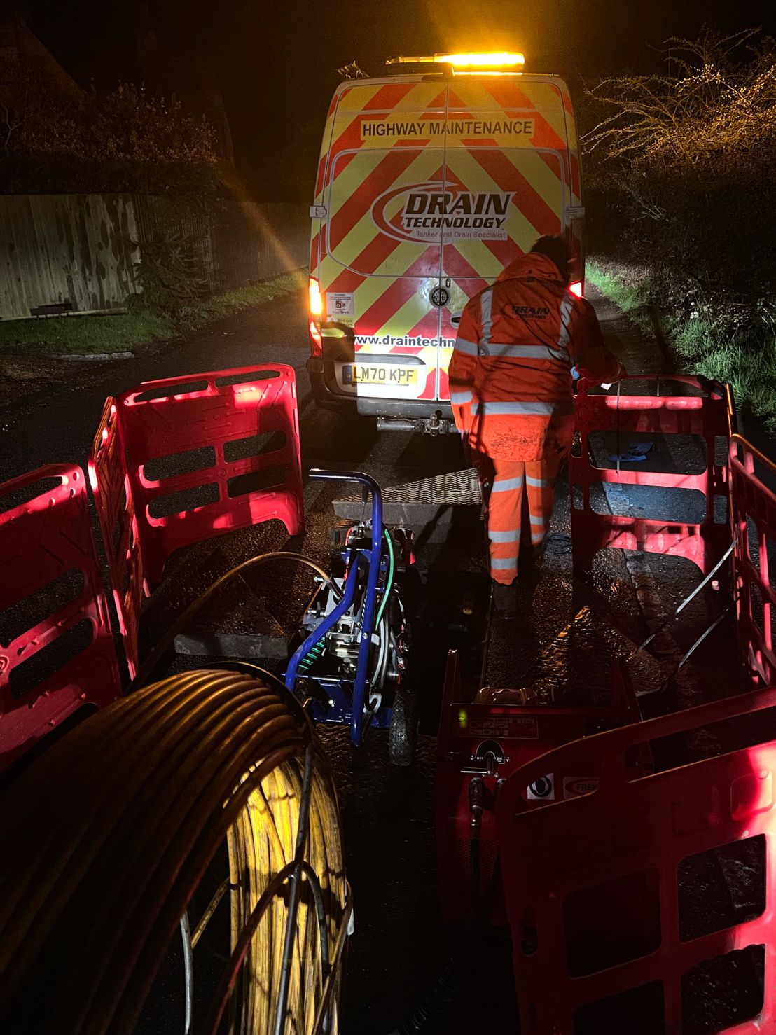 In the middle of the night, a Drain technology employee is pictured using Telecom equipment. The employee is situated behind a Highway Maintenance Drain technology van on the roadside, wearing hi-vis.