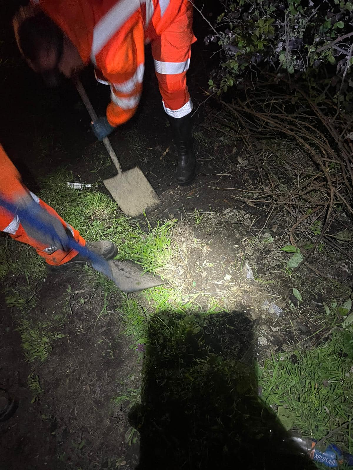 Working onsite at night, the lower extremity of two employees wearing orange hi-vis are pictured shovelling soil on a grassy, muddy field.