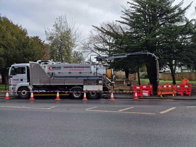the side profile of a Tanker Drain Technology vehicle is the main subject of the image. it is located on the side of a road which has been coned off. At the rear of the vehicle, pipework is coming out from the top end and going into the path drain where the tanker is parked. This area is fenced off and a employee wearing hi-vis is standing at the back.