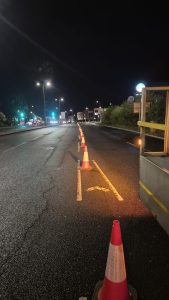 Four lane road in darkness being lit by street lamps. Traffic cones separate one lane from the rest. 