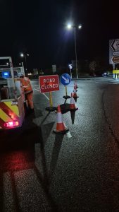 Road closure sign and traffic cones closing an exit on a roundabout. Highway maintenance van behind the cones. 