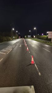 Four lane road at night, lit up by streetlights with two lanes separated by a line of cones. 
