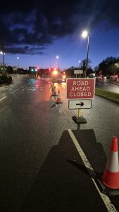 "Road Ahead Closed" sign leading up to a roundabout at night time, lit up by street lights. One lane closed with traffic cones and a highway maintenance vehicle. 