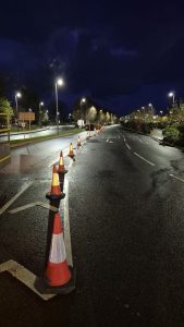 Four lane road at night, lit up by streetlights with two lanes separated by a line of cones. 
