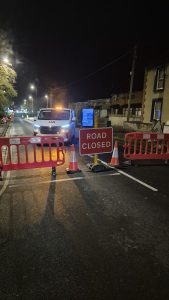 Closed road at night time  lit up by streetlights. Road closed sign in the centre with a traffic cone either side and barriers.