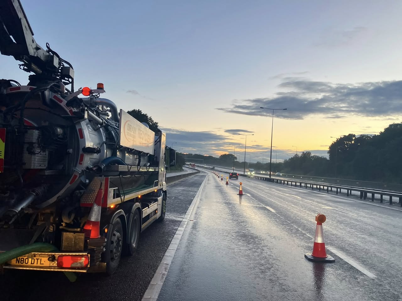 Drain Technology Service truck parked on wet highway at sunrise, traffic cones closing a lane and cars in the distance.