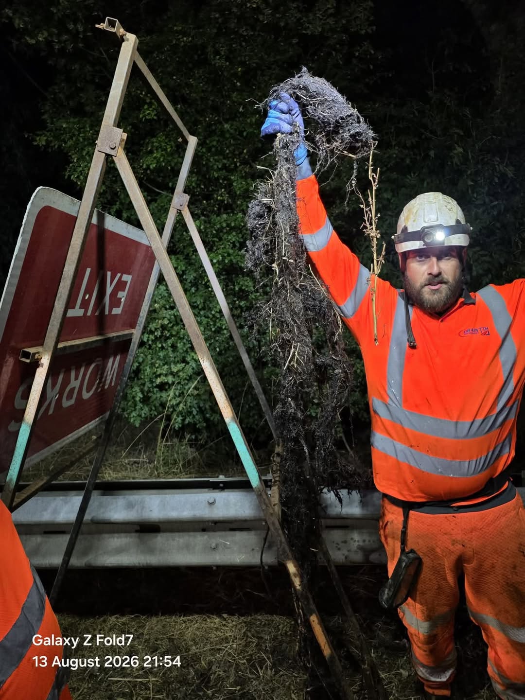 Man in orange hi vis holding a root nexxt to a temporary road sign