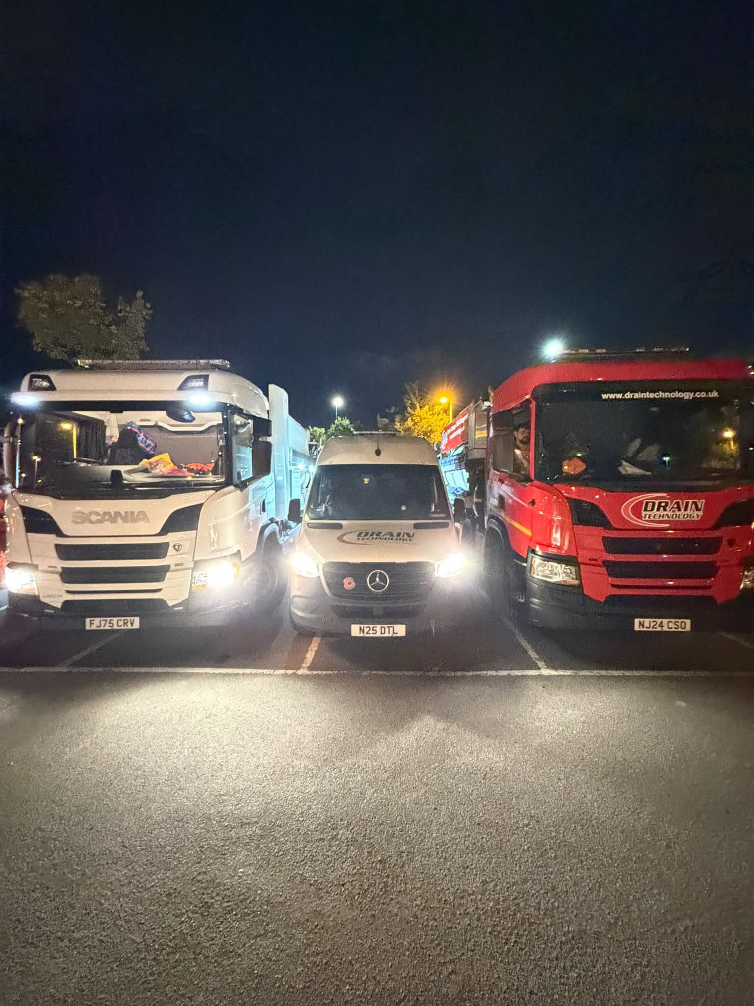 a white lorry with its lights on, a white van with its lights on and a red lorry with its lights on in a dark car park
