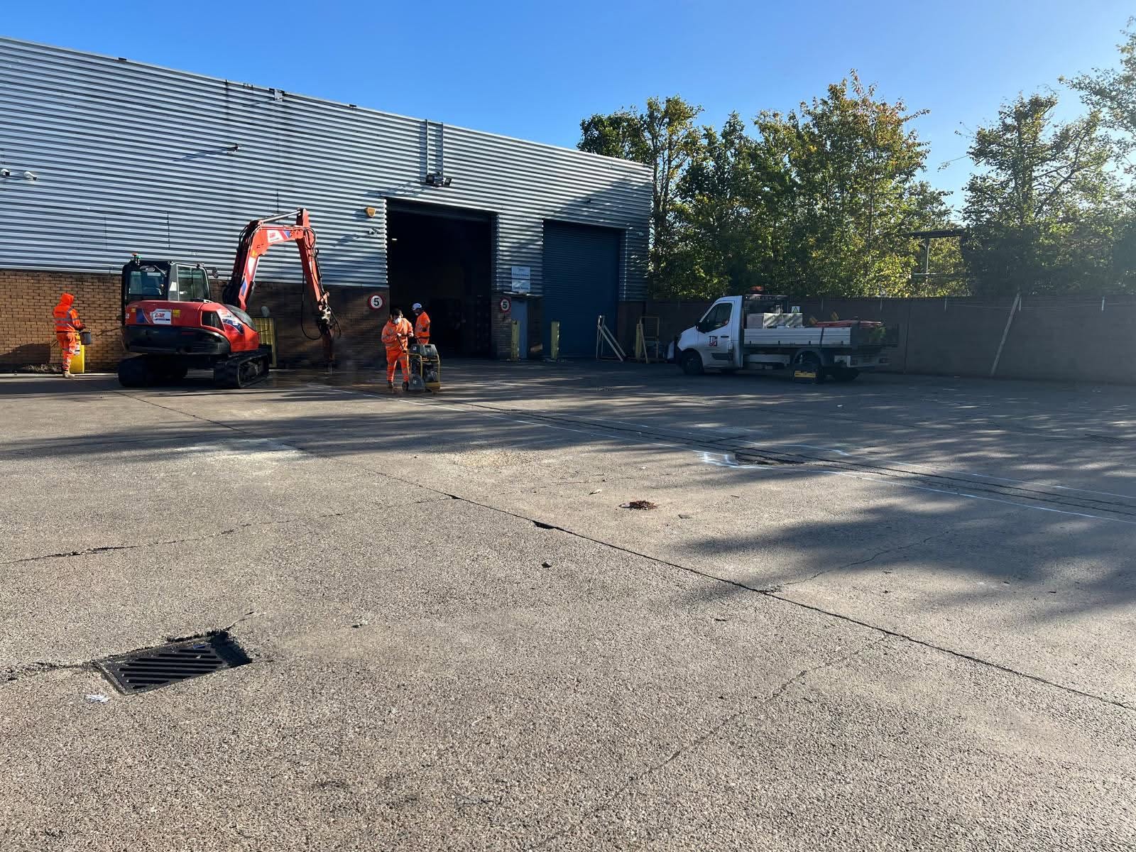 workers wearing orange working in a carpark by a large warehouse with a digger and a truck